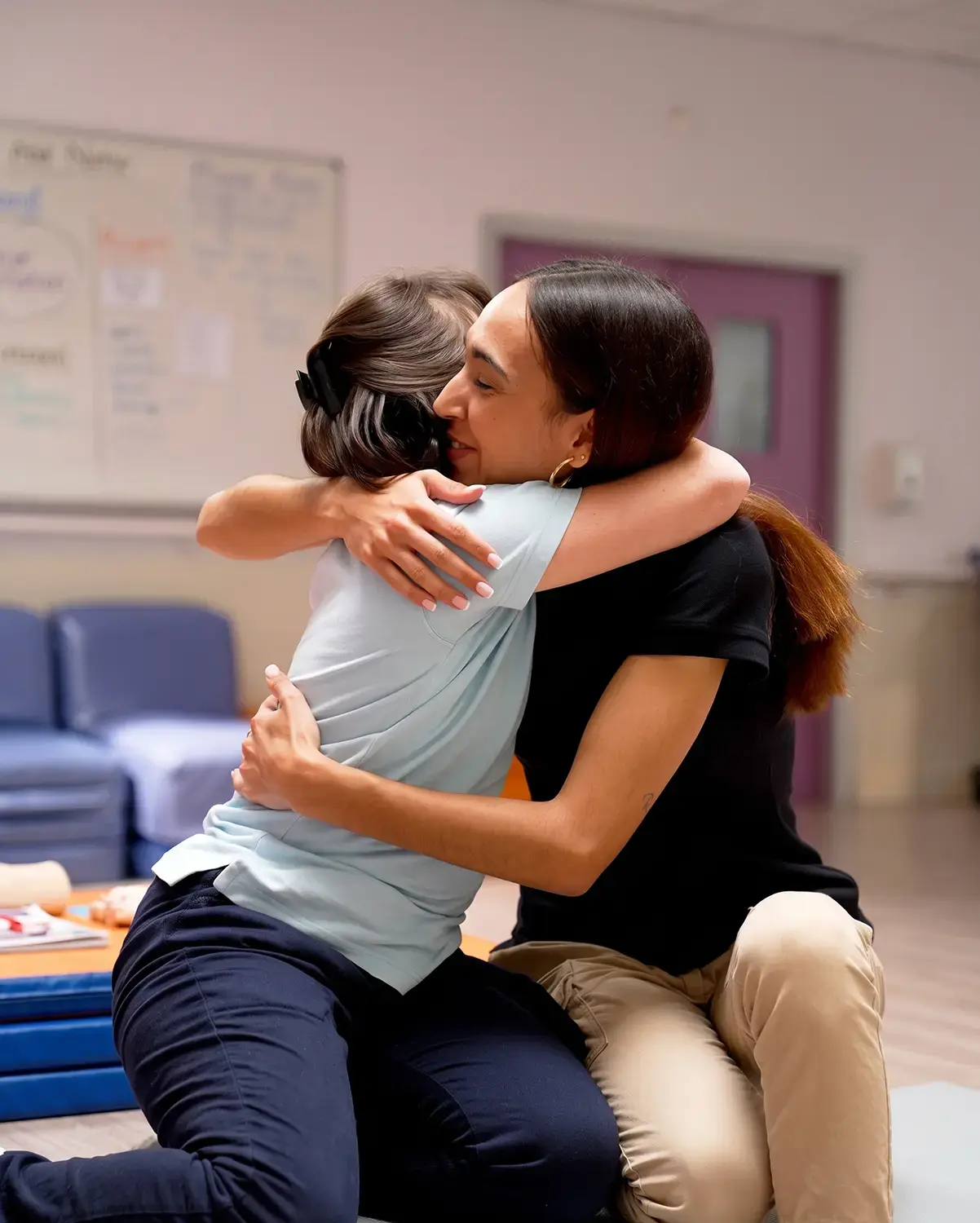 Dos jóvenes, una con uniforme y otra con ropa casual, se abrazan en un gesto de apoyo emocional y psicosocial en el interior de un centro socioeducativo o de atención